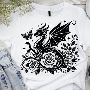 Puede incluir: Dise&ntilde;o gr&aacute;fico en blanco y negro de un drag&oacute;n con las alas extendidas, rodeado de rosas y una mariposa. El dise&ntilde;o est&aacute; en una camiseta blanca.