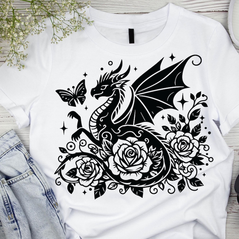 Puede incluir: Dise&ntilde;o gr&aacute;fico en blanco y negro de un drag&oacute;n con las alas extendidas, rodeado de rosas y una mariposa. El dise&ntilde;o est&aacute; en una camiseta blanca.