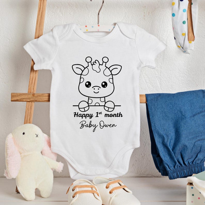 May include: White baby onesie with a black and white giraffe graphic. The text "Happy 1st month Baby Owen" is printed below the giraffe. The onesie is hanging on a hanger with other baby items in the background.