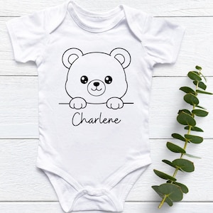 May include: White baby onesie with a black and white bear graphic. The bear is peeking over a line with the name "Charlene" written in cursive below. The onesie has short sleeves and a snap closure.