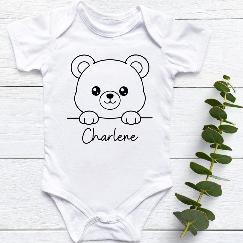 May include: White baby onesie with a black and white bear graphic. The bear is peeking over a line with the name "Charlene" written in cursive below. The onesie has short sleeves and a snap closure.