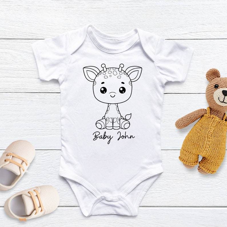 K&ouml;nnte beinhalten: Wei&szlig;er Baby-Body mit einer schwarz-wei&szlig;en Giraffen-Grafik und dem Namen "Baby John" in Schreibschrift. Ein beiger Teddyb&auml;r und Babyschuhe liegen neben dem Body. Der Body ist aus weichem Material.