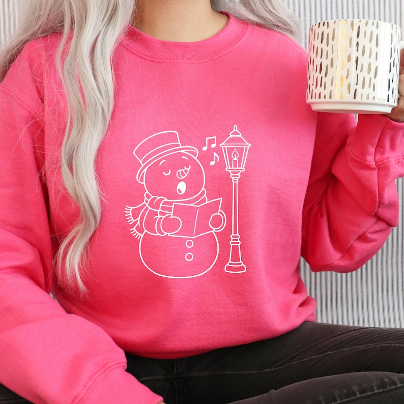 May include: A bright pink sweatshirt featuring a white line drawing of a singing snowman wearing a hat and scarf, with a street lamp and musical notes. The person is holding a white mug with gold vertical stripes.