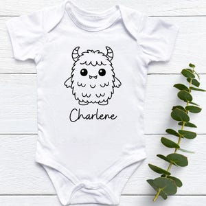 May include: White baby onesie with a black and white cartoon monster design and the name "Charlene" in a script font. The monster has horns, large eyes, and a fluffy body. The onesie is short-sleeved.