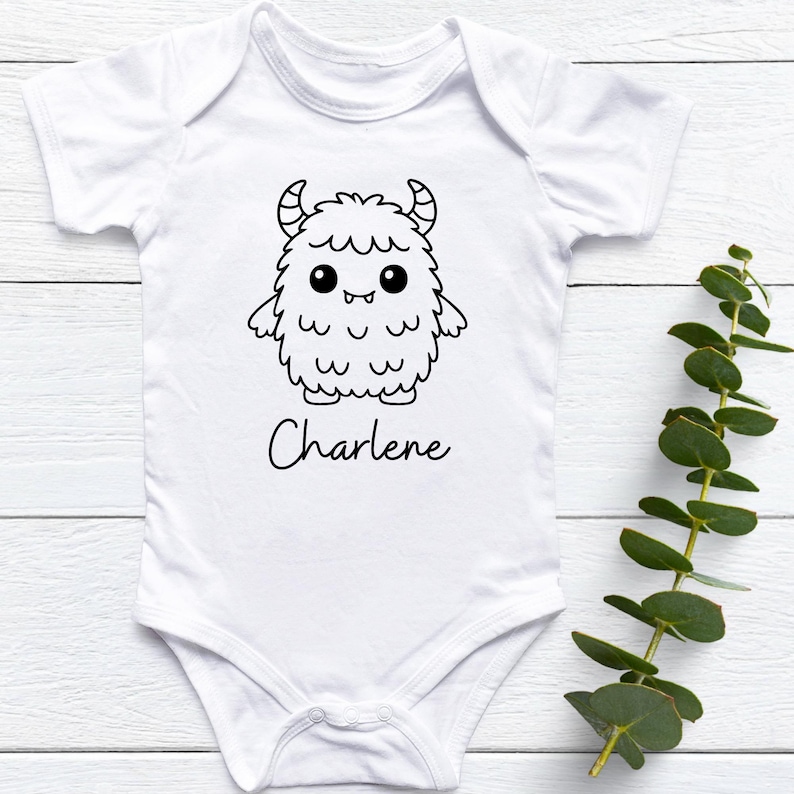 May include: White baby onesie with a black and white cartoon monster design and the name "Charlene" in a script font. The monster has horns, large eyes, and a fluffy body. The onesie is short-sleeved.