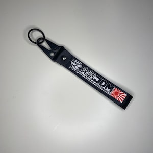 Peut inclure: Porte-clés noir avec mousqueton et anneau noirs. La sangle du porte-clés présente du texte japonais blanc et un motif de soleil levant sur fond noir. Le texte se lit "OSAKA-KANJO-PERFORMANCE" et "JDM".