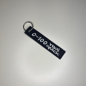 Pode incluir: Chaveiro retangular preto com texto bordado branco que diz "0-100 real quick". O chaveiro tem um anel de metal prateado na parte superior. O fundo é uma superfície branca lisa.