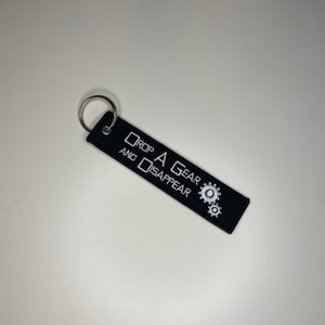 May include: Black keychain tag with white embroidered text that reads "DROP A GEAR AND DISAPPEAR." The tag features two white gear graphics and a silver key ring. The tag is rectangular and made of a woven material.
