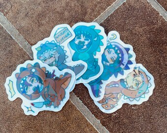 Pode incluir: Uma coleção de autocolantes holográficos com personagens de desenhos animados vestidos com fantasias de dinossauros. Um autocolante tem o texto "NAUGHTY CORNER". Os autocolantes são em tons de azul, castanho e verde.