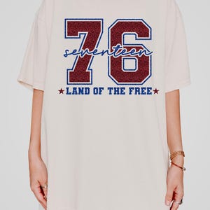 May include: An off-white short-sleeve t-shirt featuring a large graphic with the number 76 in dark red glitter with a blue outline. The word seventeen is written in blue script across the numbers, with the text LAND OF THE FREE below.