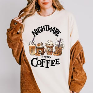 May include: A cream-colored crewneck sweatshirt featuring a graphic print of four coffee cups with Halloween-themed designs, including pumpkins and character motifs. The text "NIGHTMARE BEFORE COFFEE" is printed in black above and below the cups.