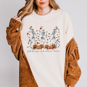 May include: A cream-colored crewneck sweatshirt featuring a graphic of three dancing skeletons surrounded by orange pumpkins, sunflowers, and falling autumn leaves. The text "fall breeze and autumn leaves" is printed in black cursive below the graphic.