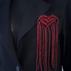 May include: A black blazer featuring a red heart-shaped decoration. The heart is crafted from shimmering red strands that hang down the front of the jacket. The blazer has a traditional notched lapel and a single-button fastening.