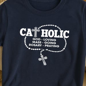 May include: Navy blue t-shirt with the word "CATHOLIC" in white, above a rosary design and the words "GOD - LOVING MASS - GOING ROSARY - PRAYING". A cross is at the top of the word and at the end of the rosary.