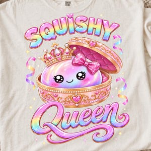May include: An off-white t-shirt featuring a graphic of a round, iridescent, pink character with large black eyes and a pink bow, wearing a gold crown inside an open, ornate pink jewelry box. The text SQUISHY QUEEN is written in rainbow gradient.