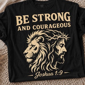 May include: A black short-sleeve t-shirt featuring a beige graphic print of a lion and a person wearing a crown of thorns. The text reads BE STRONG AND COURAGEOUS and Joshua 1:9. The design is centered on the front of the garment.