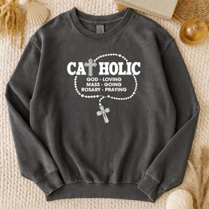 May include: Dark gray sweatshirt with the word "CATHOLIC" in white, featuring a cross design. Below the word are the phrases "GOD - LOVING", "MASS - GOING", "ROSARY - PRAYING" in white. A white rosary with a cross hangs below the text.