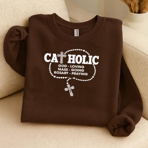 May include: A brown sweatshirt with the word "CATHOLIC" in white, along with a cross and rosary design. Below the word "CATHOLIC" are the words "GOD - LOVING, MASS - GOING, ROSARY - PRAYING". The sweatshirt is folded on a beige surface.
