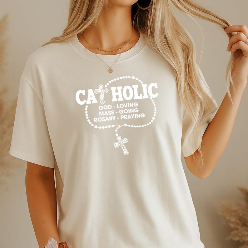 May include: Cream-colored t-shirt with the word "CATHOLIC" in white, featuring a cross design. Below the word are the words "GOD - LOVING, MASS - GOING, ROSARY - PRAYING". A rosary with a cross is also featured.
