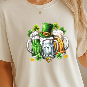 May include: Cream-colored t-shirt featuring a St. Patrick's Day design. The graphic includes three beer mugs with green, white, and amber-colored beer, a green top hat, shamrocks, gold coins, and a horseshoe.