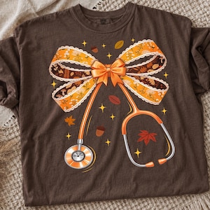 Puede incluir: Una camiseta de color marrón oscuro con un estampado gráfico de un estetoscopio naranja integrado en un lazo decorativo grande. El lazo tiene un patrón otoñal en naranja y marrón con hojas, setas y bellotas, acentuado por pequeñas estrellas amarillas.