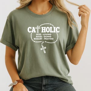 May include: Olive green t-shirt with the word "CATHOLIC" in white, above a rosary design and the words "GOD - LOVING, MASS - GOING, ROSARY - PRAYING". The shirt has short sleeves and a crew neck.