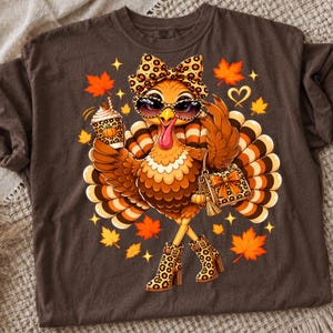 Può includere: Una t-shirt marrone scuro con la grafica di un tacchino che indossa occhiali da sole, un fiocco e stivali con stampa leopardata. Il tacchino tiene in mano una tazza di caffè e una borsa, circondato da foglie d'acero arancioni e piccole stelle gialle.