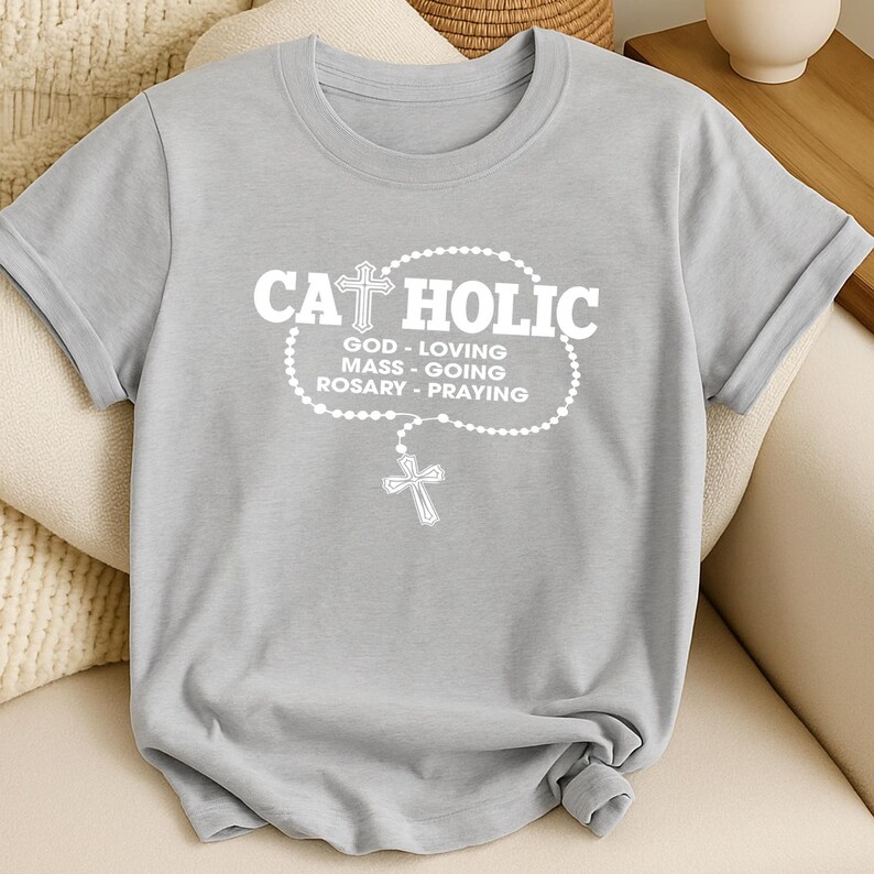 May include: A light gray t-shirt with the word "CATHOLIC" in white, along with a rosary design and the words "GOD - LOVING, MASS - GOING, ROSARY - PRAYING". The shirt has short sleeves and a crew neck.