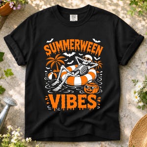 Peut inclure: Un t-shirt noir avec un motif représentant un squelette portant des lunettes de soleil, se relaxant dans une bouée rayée orange et blanche. Le design inclut des palmiers, des chauves-souris, un ballon de plage, un seau citrouille et le texte SUMMERWEEN VIBES.