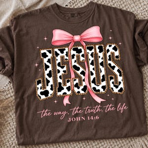May include: A brown t-shirt featuring the word JESUS in a black and white cow print pattern with a leopard print border. A large pink bow sits atop the letters. Below, the text reads: the way, the truth, the life, JOHN 14:6 in pink script.
