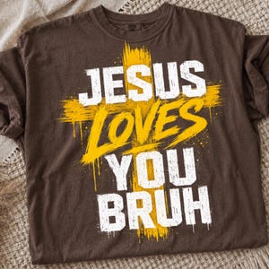 May include: A dark brown short-sleeve t-shirt featuring a large yellow cross graphic behind the white distressed text that reads JESUS LOVES YOU BRUH. The design has a paint-splatter effect with drips extending from the cross and the lettering.