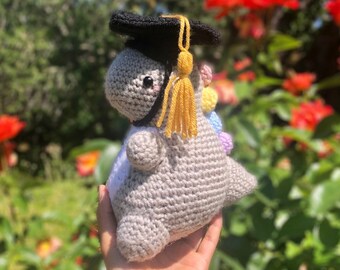 May include: A gray crocheted dinosaur wearing a black graduation cap with a yellow tassel. The dinosaur has a rainbow of colors on its back.