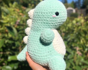 May include: A crocheted green and white dinosaur plush toy with a cute face. The dinosaur is sitting in a person's hand.