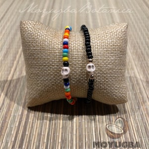 May include: Two beaded bracelets rest on a burlap pillow. One bracelet features colorful beads and a white skull charm. The other bracelet is made of black beads with a white skull charm. The image includes the text "Moyugba Botanica" and "MOYUGBÁ".