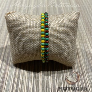 May include: Two beaded bracelets, one with green, yellow, and red beads, and the other with red and brown beads. The bracelets are displayed on a small, square, beige pillow with the text "Moyugba Botanica" and "Moyugba" in the image.