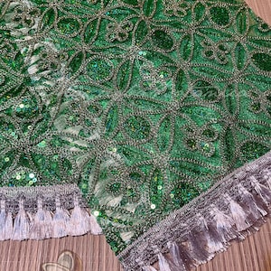 May include: Close-up of a green fabric with intricate floral embroidery and silver accents. The fabric is embellished with sequins and features a silver tassel trim along the edges. The design is detailed and ornate.