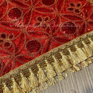 May include: A red textile with a gold floral pattern and gold sequin accents. The edge is trimmed with a gold tassel fringe. The text "Moyugba Botanica" is visible on the textile.