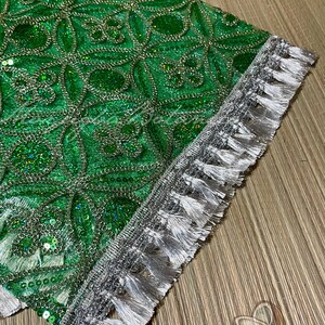 May include: Close-up of a section of green fabric with a detailed pattern of sequins and silver thread. The edge is trimmed with a silver border and white tassels, creating a decorative textile.