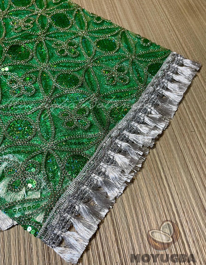 May include: Close-up of a section of green fabric with a detailed pattern of sequins and silver thread. The edge is trimmed with a silver border and white tassels, creating a decorative textile.