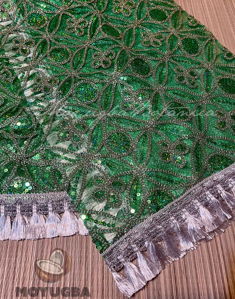 May include: Close-up of a green fabric with intricate floral embroidery and silver accents. The fabric is embellished with sequins and features a silver tassel trim along the edges. The design is detailed and ornate.