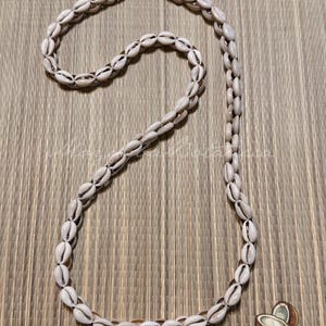 May include: A long necklace composed of numerous small, off-white cowrie shells strung together in a continuous loop. The shells are arranged in a single strand, showcasing their natural oval shapes and characteristic central slits in a light tan color.