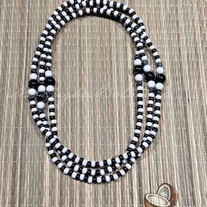May include: A layered necklace composed of black and white beads. The beads are arranged in a repeating pattern of small white beads, followed by larger black beads. The necklace is displayed on a light brown woven surface.