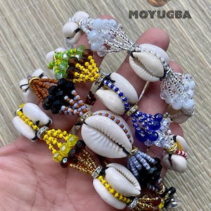 May include: A set of six colorful beaded bracelets with cowrie shells. Each bracelet features a different color scheme and bead pattern, including yellow, blue, red, white, and black beads. The bracelets are adorned with small, clear crystals and have a tassel at the end.