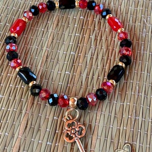 May include: A beaded bracelet featuring alternating black and red faceted beads, interspersed with small gold-toned spacers. A gold-toned metal key charm hangs from the bottom of the bracelet. The text @MOYUGBABOTANICA is visible at the top and MOYUGBA at the bottom.