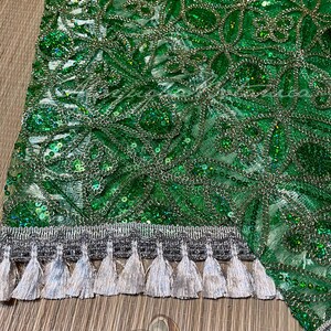 May include: Close-up of a green fabric with intricate floral patterns, sequins, and silver thread detailing. The bottom edge features a silver trim with hanging tassels. The fabric has a shimmering, textured appearance.