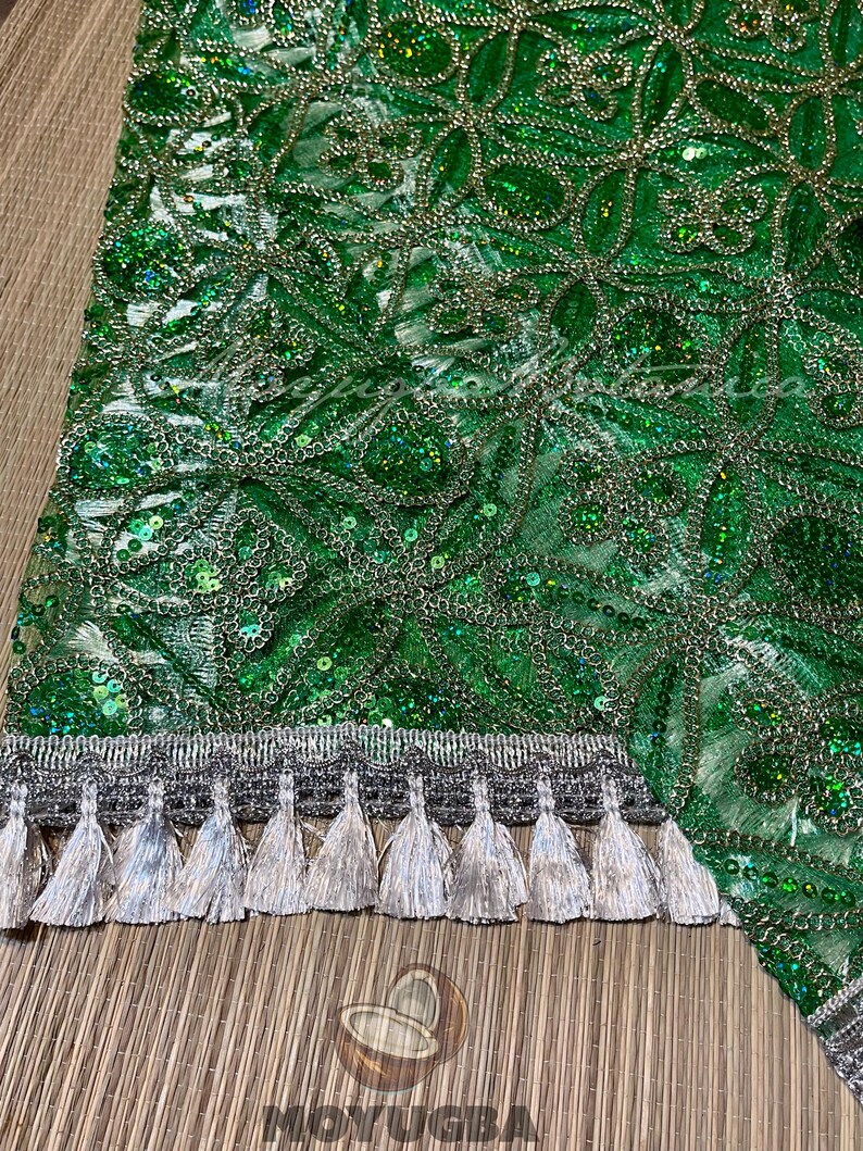 May include: Close-up of a green fabric with intricate floral patterns, sequins, and silver thread detailing. The bottom edge features a silver trim with hanging tassels. The fabric has a shimmering, textured appearance.