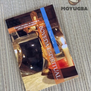 May include: A book titled "BOVEDA ESPIRITUAL" by Oishavn Ata Erinde. The cover features a dark wooden cross with a gold trim, a dark glass vase, and a lit candle. The book cover is a light blue color with white text.
