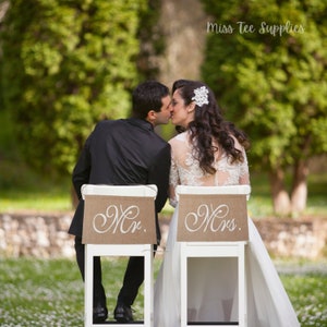 Op de afbeelding: Een bruid en bruidegom kussen elkaar terwijl ze op witte stoelen zitten met jute stoelbanden waarop in witte cursieve letters "Mr." en "Mrs." staat. Het paar bevindt zich in een tuinsetting met groene bomen en witte bloemen op de achtergrond.