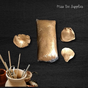 May include: A bag of gold mica powder, along with three gold mica flakes. The mica powder is in a clear plastic bag and the flakes are on a black surface. Miss Tee Supplies is written in white text on a black background.