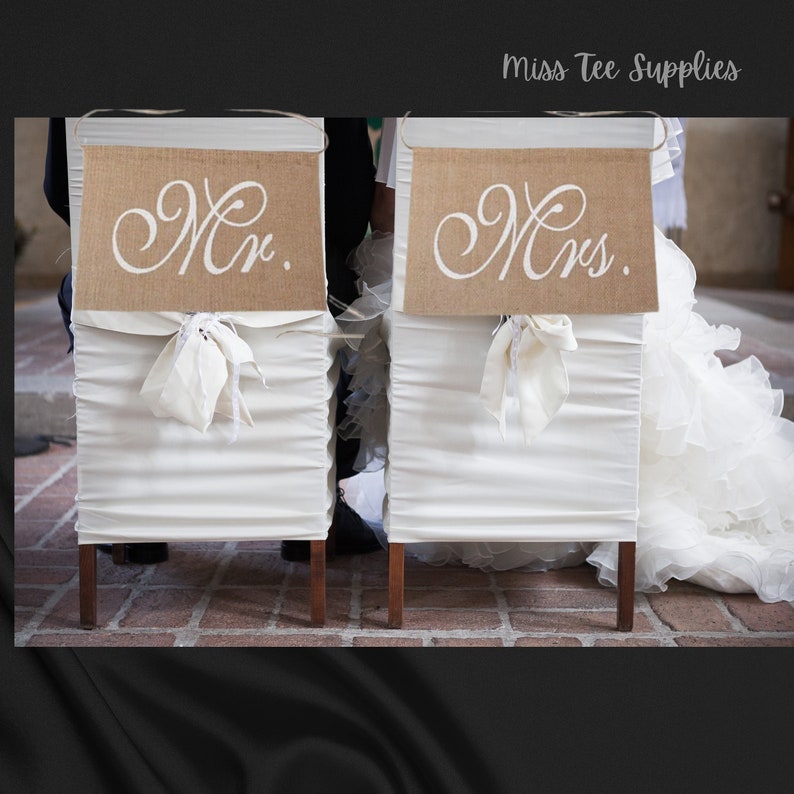 Op de afbeelding: Twee witte stoelen met juteborden waarop 'Mr.' en 'Mrs.' staat. De stoelen zijn opgesteld voor een huwelijksceremonie.